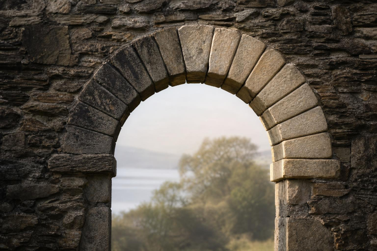 Dokumentarisch-realistische Fotografie eines alten keltischen Rundbogens aus unregelmäßigen Natursteinen in einer historischen irischen Kirchenruine aus dem 11. Jahrhundert. Der Bogen zeigt oben in der Mitte einen deutlichen Schlussstein, rechts und links jeweils sieben hellere bzw. dunklere Steine. Durch die Öffnung fällt weiches Morgenlicht, im Hintergrund unscharfe Bäume vor einem See.