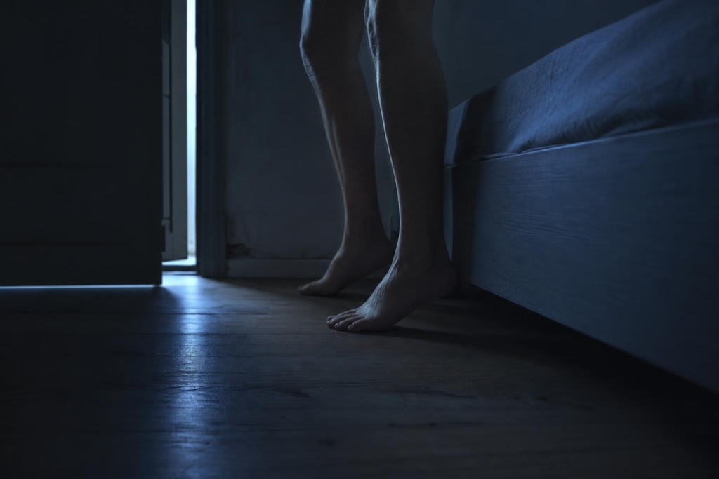 Dokumentarische Fotografie eines dunklen Schlafzimmers bei Nacht, niedrige Perspektive knapp über dem Boden, scharf konturierte Bettkante im Vordergrund, zwei nackte Füße halten vorsichtig inne, schmaler Streifen kühlen Lichts fällt durch halb geöffnete Tür auf Boden und Bettkante, gedeckte Blau- und Grautöne, ruhige Atmosphäre.